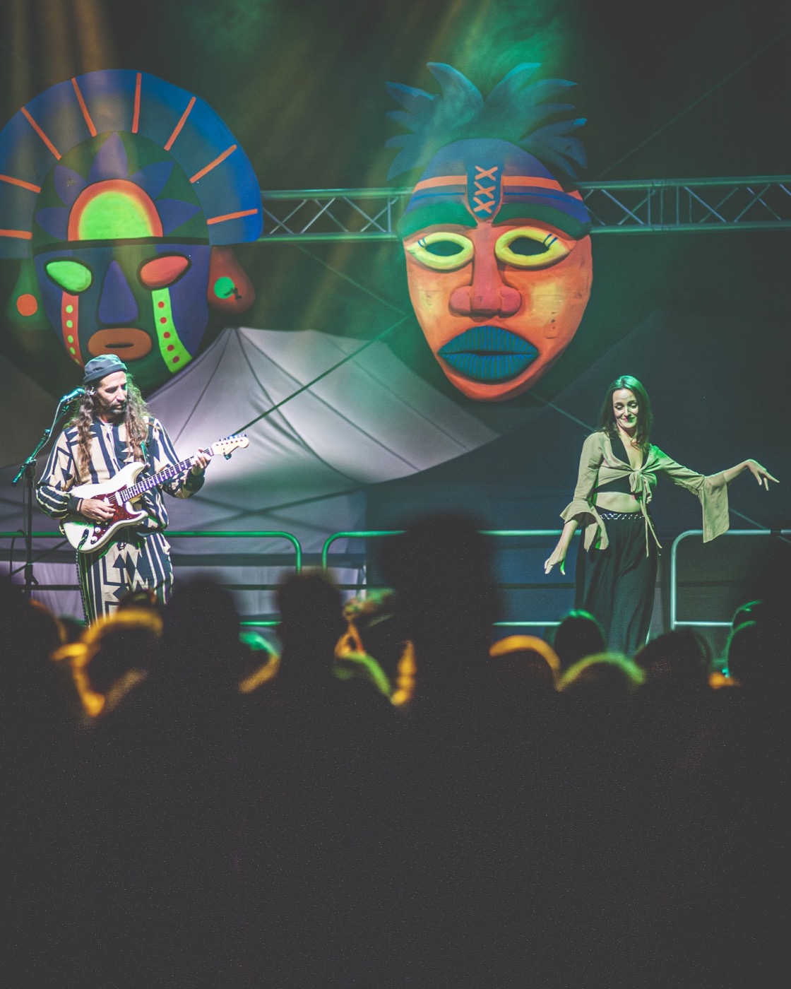 Wiktoria performing a dance on stage next to a guitarist during a concert, colourful masks behind them